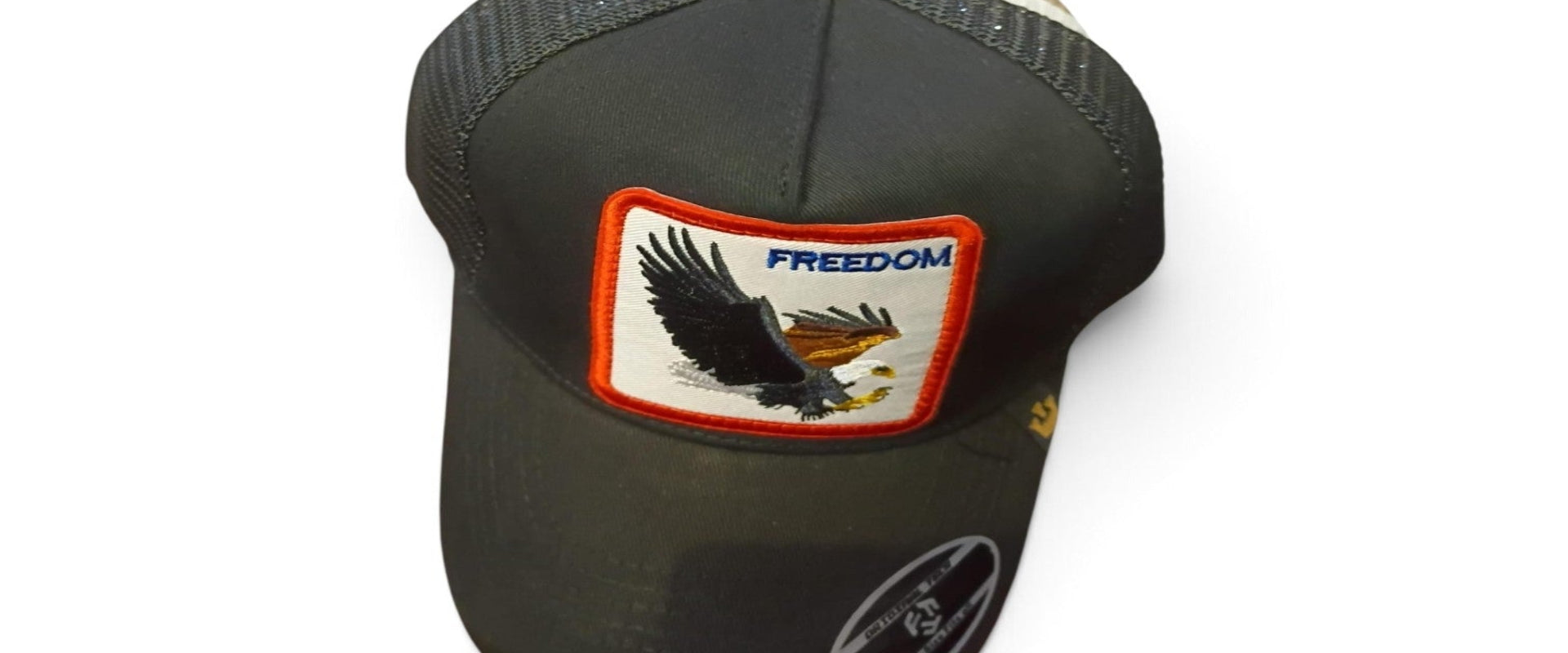 A black trucker-style cap with a mesh back, featuring a rectangular embroidered patch on the front with an eagle design and the word 'FREEDOM' in blue. A circular sticker is visible on the brim.